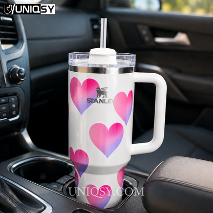 A white Stanley travel tumbler with a handle, clear lid, and straw, decorated with pink and purple gradient hearts, shown sitting in a car cup holder. The Stanley logo is printed on the front.