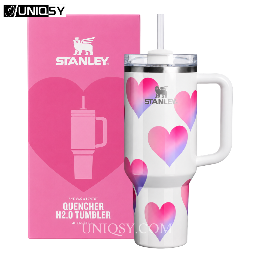 A white Stanley Quencher H2.0 tumbler with pink and purple heart patterns, shown next to its pink retail box on a white background.