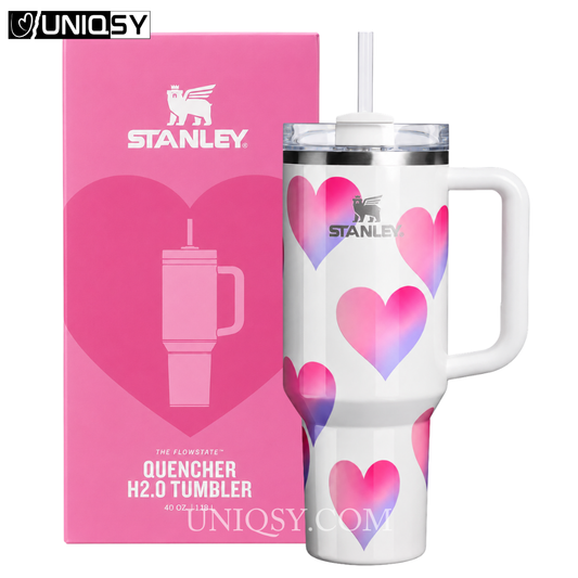A white Stanley Quencher H2.0 tumbler with pink and purple heart patterns, shown next to its pink retail box on a white background.