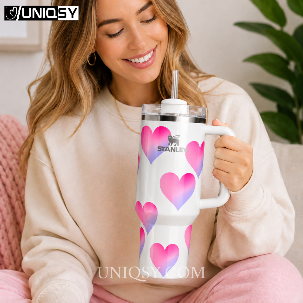 A woman in a cream sweatshirt holds a white Stanley tumbler with a handle and clear straw. The tumbler is decorated with pink and purple heart patterns, and the brand name Stanley is printed on the front.