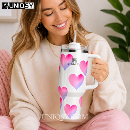 A woman in a cream sweatshirt holds a white Stanley tumbler with a handle and clear straw. The tumbler is decorated with pink and purple heart patterns, and the brand name Stanley is printed on the front.