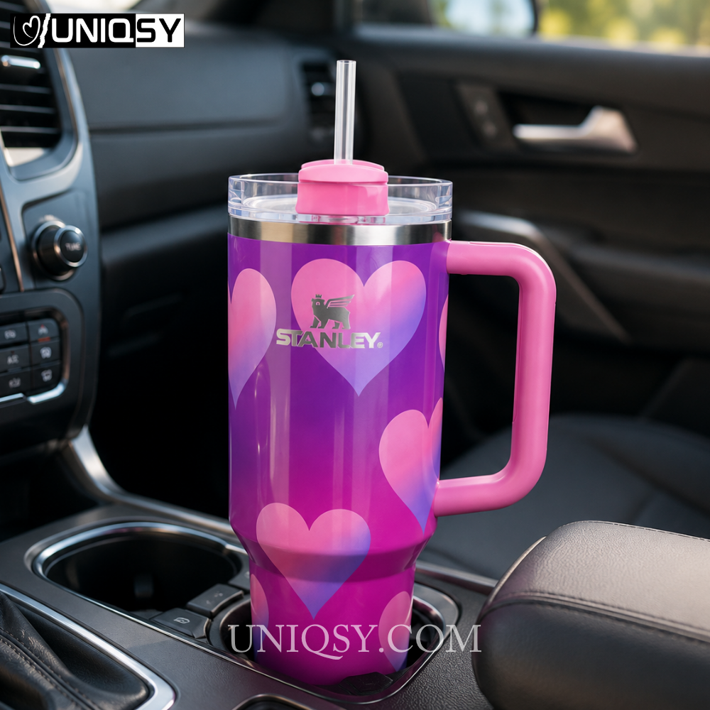 A Stanley insulated tumbler with a pink handle and lid, featuring a purple and pink heart pattern, shown sitting in a car cup holder.