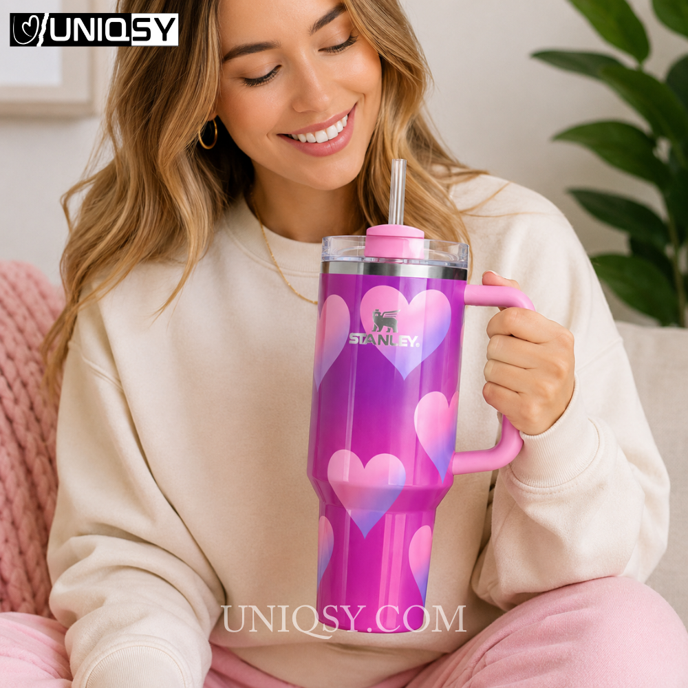 A woman in a cream sweatshirt holds a large pink and purple Stanley tumbler with heart patterns and a clear straw. The image has a UNIQSY watermark in the top left and bottom center.