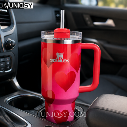 A red and pink Stanley insulated tumbler with heart patterns, a red handle, and a clear lid with a straw, shown sitting in a car cup holder.